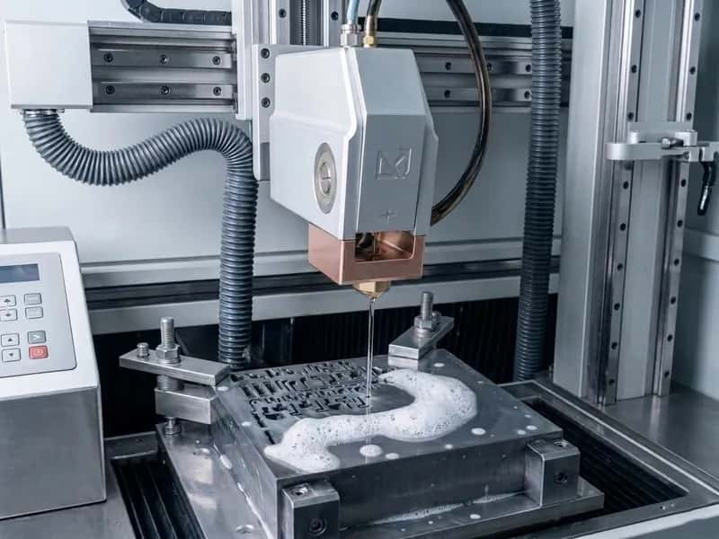 Wire EDM machine head cutting a clamped steel mold block with dielectric foam on the surface